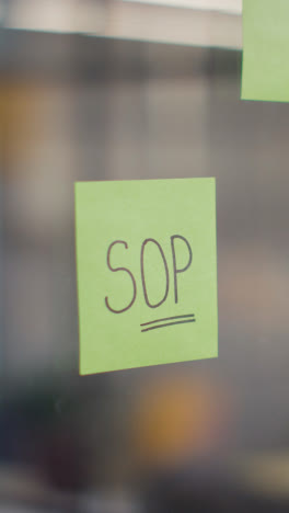 Vertical-Video-Close-Up-Of-Woman-Putting-Sticky-Note-With-SOP-Written-On-It-Onto-Transparent-Screen-In-Office